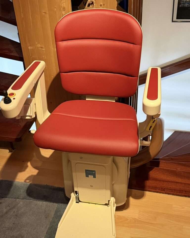 Projekte Ein rot-weißer Paitsch Freecurve Kurventreppenlift, frontal fotografiert in einem Wohnhaus in 71282 Hemmingen. Gut zu sehen sind der ergonomische rote Sitz, die Steuerungselemente an den Armlehnen und die ausgeklappte Fußstütze auf dem Holzfußboden.