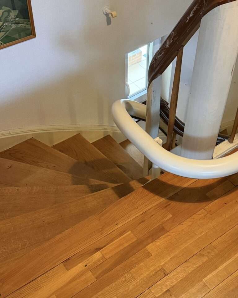Projekte Blick von oben auf eine gewendelte Holztreppe mit der sauber installierten, geschlossenen Schiene des Treppenlifts, die eng am Geländer entlangführt.