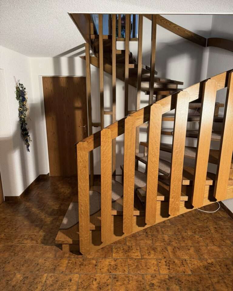 Projekte Gesamtansicht einer offenen, gewendelten Holztreppe mit markantem Geländer im Erdgeschoss eines Wohnhauses vor der Installation des Treppenlifts von Paitsch Lifte.