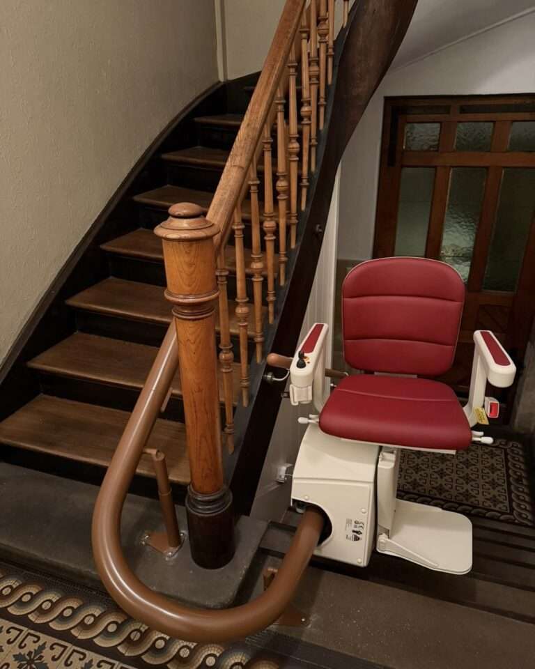 Projekte Ein roter Paitsch Kurven-Treppenlift für mehrere Etagen, montiert an einer klassischen Holztreppe in Ludwigsburg. Sichtbar ist die platzsparende Schienenführung mit einer automatischen Klappschiene im Startbereich des Treppenhauses.