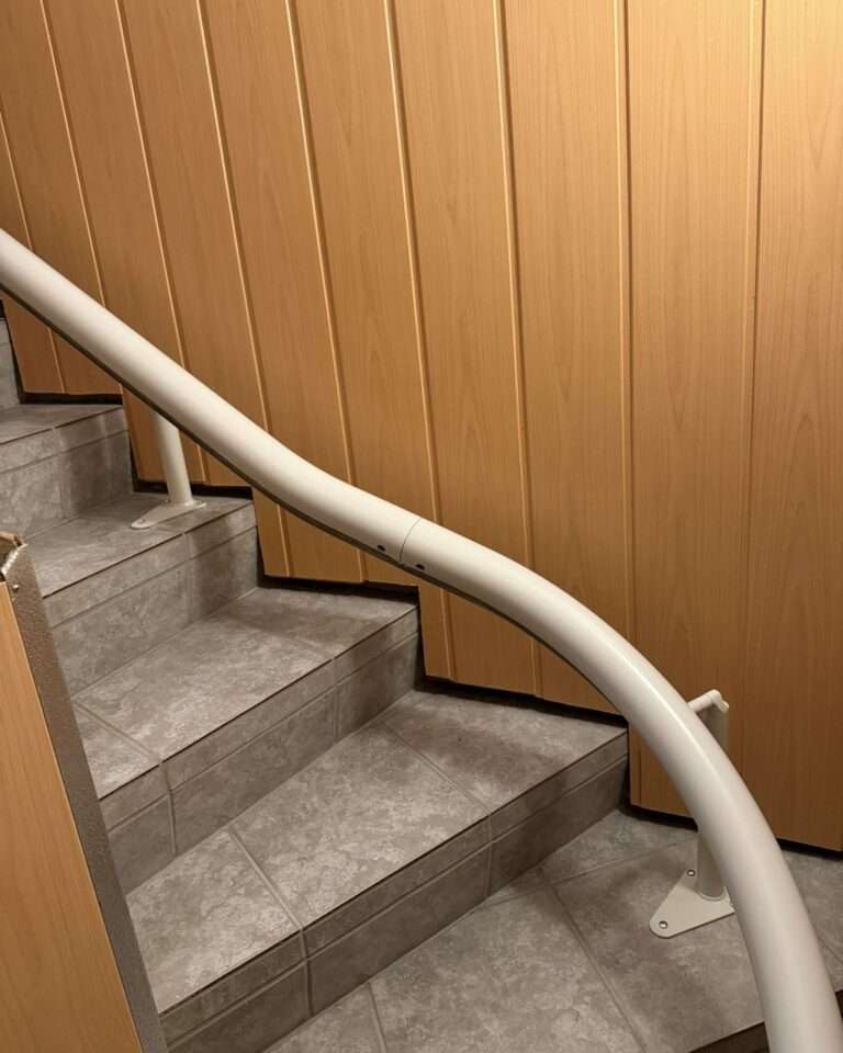Projekte Detailansicht der passgenauen weißen Einrohr-Führungsschiene des Kurventreppenlifts an einer Wandverkleidung in Holzoptik.