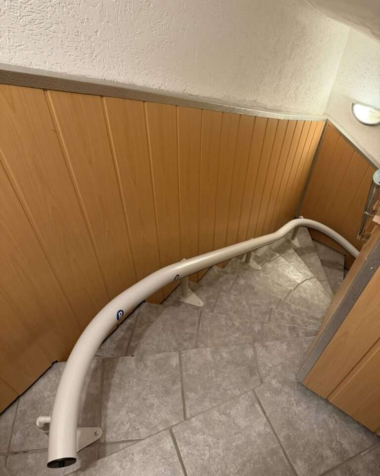 Projekte Perspektive von oben auf die steile, gewendelte Treppe mit installierter weißer Liftschiene entlang der Holzpaneele.