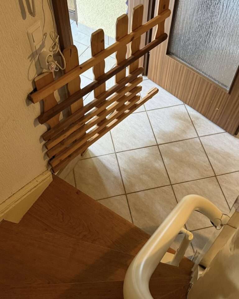 Projekte Eine geöffnete, hölzerne Absperrtür für Haustiere an einer gewendelten Holztreppe, direkt neben der weißen Schiene eines installierten Treppenlifts.