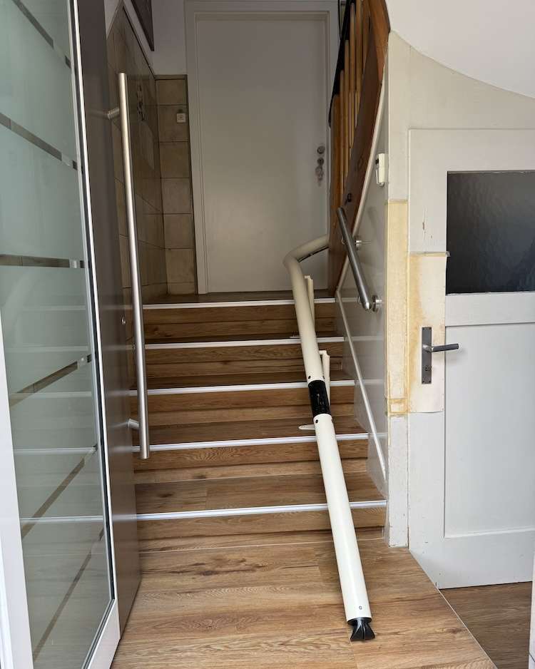 Projekte Individuelles weißes Einrohr-Schienensystem eines Treppenlifts im Hauseingang, montiert auf Holzstufen.