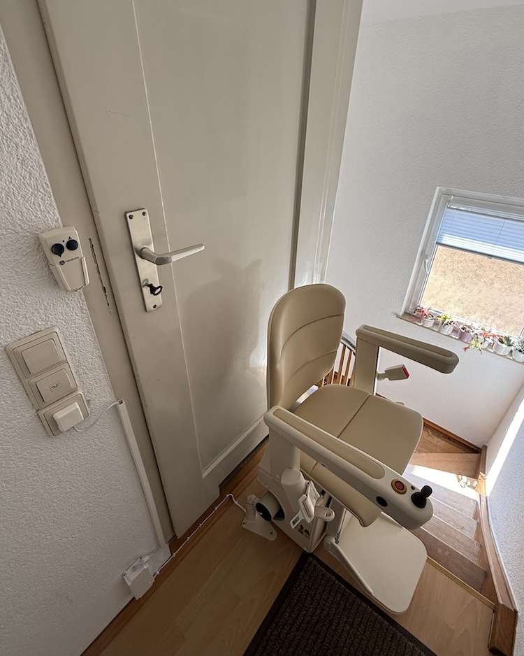 Projekte Beiger Treppenlift in oberer Parkposition direkt neben einer angepassten weißen Zimmertür im Dachgeschoss.