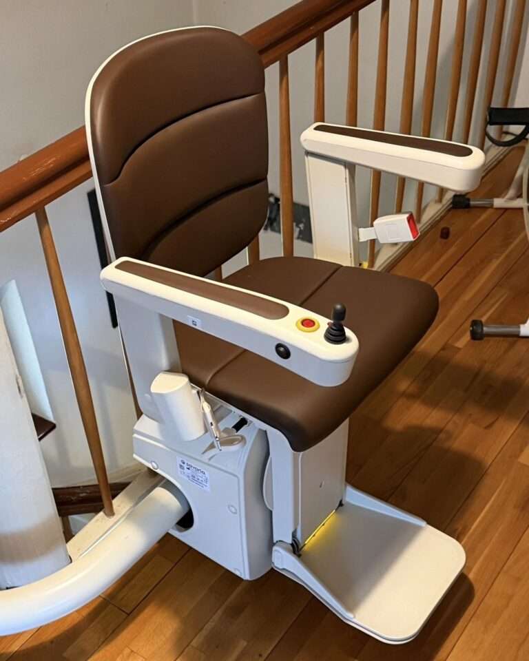 Projekte Der installierte Treppenlift mit einem komfortablen, braun gepolsterten Sitz, Armlehnen und Joystick-Bedienelement, geparkt im oberen Stockwerk an einem Holzgeländer.