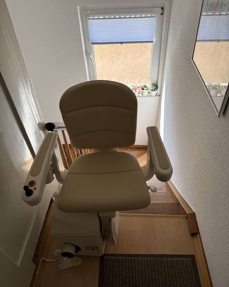 Projekte Frontansicht des beigen Treppenlift-Sitzes mit Armlehnen und Bedienelementen in der oberen Etage vor einem Fenster.