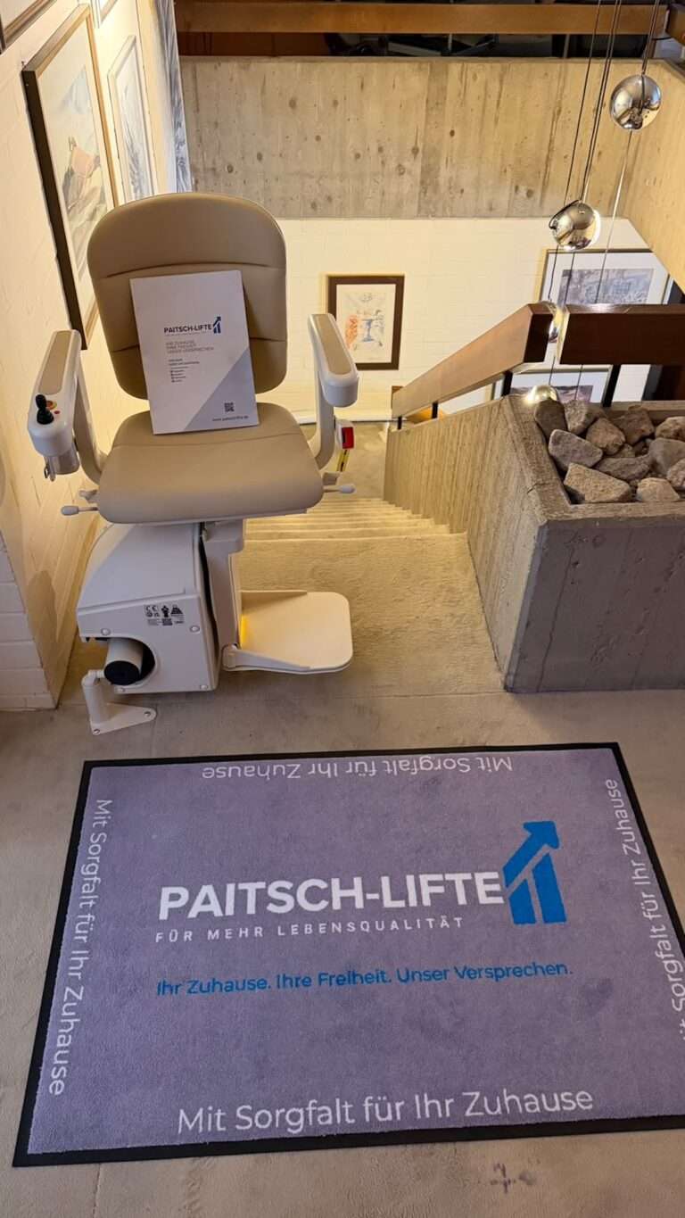 Montierter Sitzlift von Paitsch-Lifte am oberen Ende einer Treppe mit Unternehmens-Fußmatte und Infomaterial.
