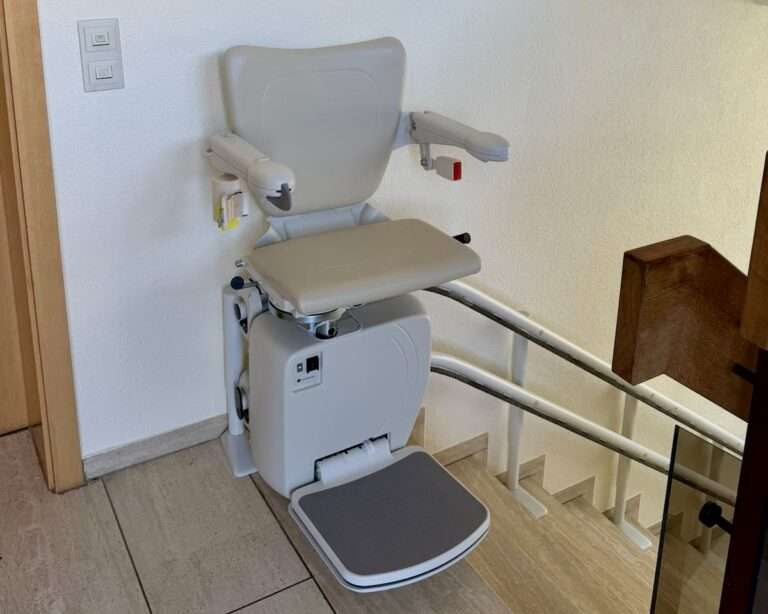 Montierter, hellgrauer Sitzlift von Paitsch-Lifte an einem Treppenaufgang mit ausgeklappter Fußstütze und Armlehnen.
